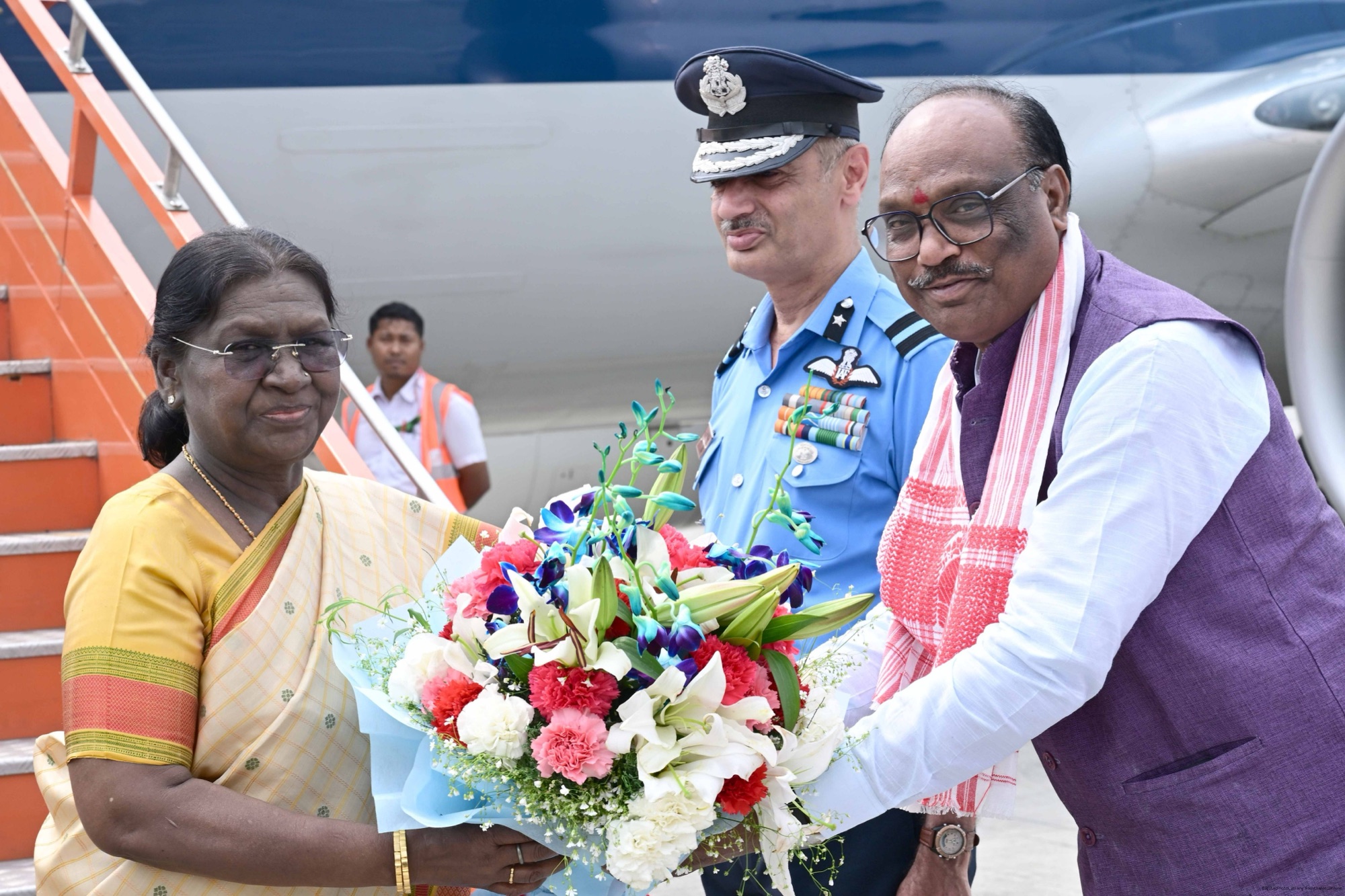 Durga Das Uikey, in a purple waistcoat and patterned stole, presenting a bouquet to President Droupadi Murmu beside an aircraft stairway, with an Indian Air Force officer standing between them
