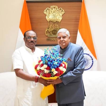 Durga Das Uikey presenting a bouquet of blue orchids and red roses to Vice-President Jagdeep Dhankhar, in front of the State Emblem and national flags