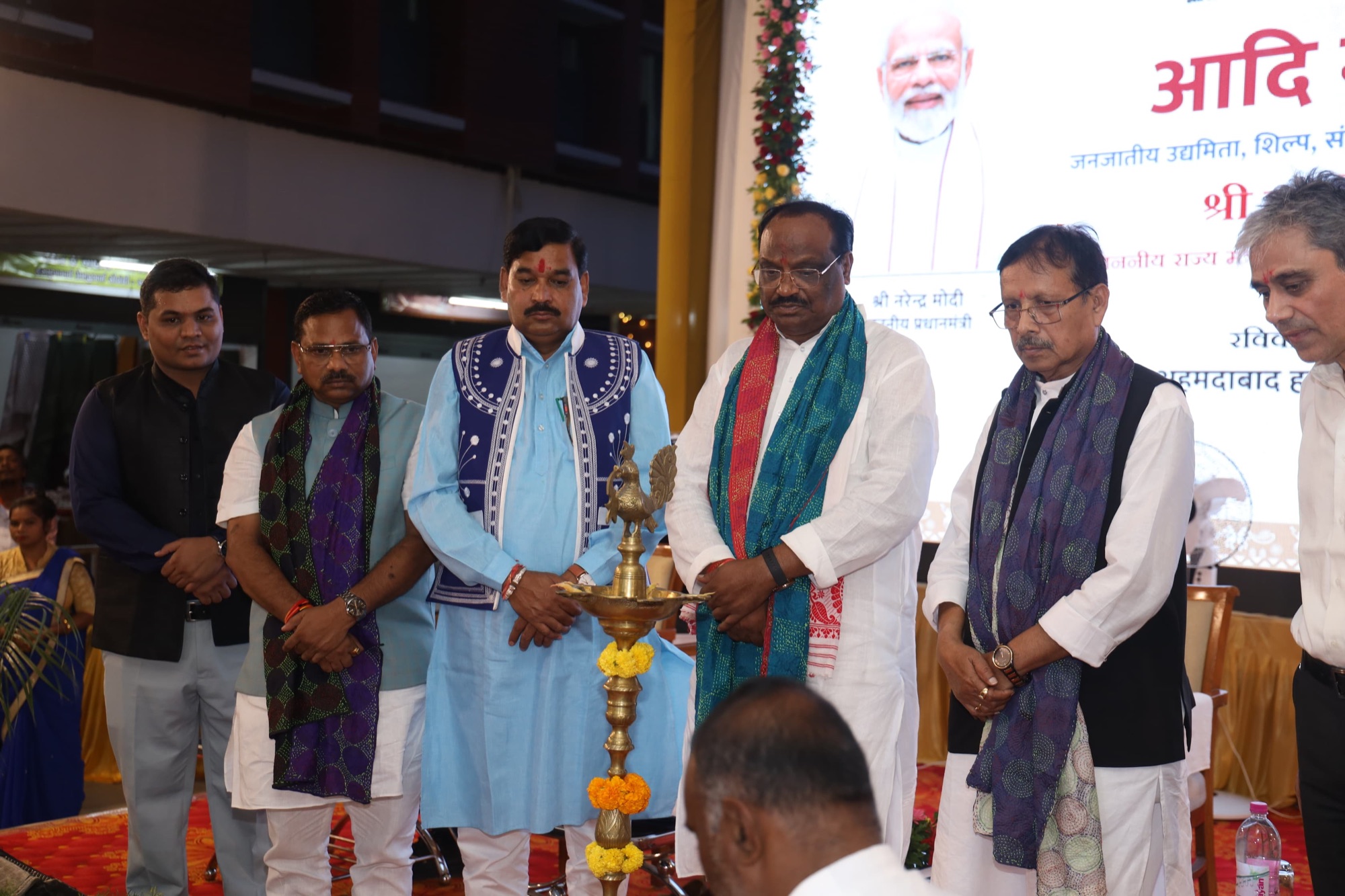 Durga Das Uikey, wearing a teal stole, standing with dignitaries beside a ceremonial brass lamp in front of a banner for Aadi Mahotsav