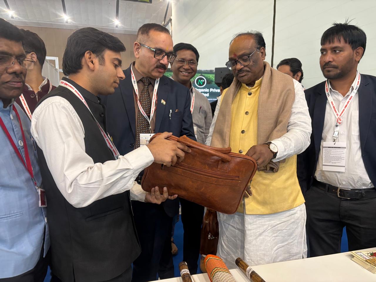 Durga Das Uikey, in a yellow kurta and beige shawl, examining a handcrafted leather bag held out by an exhibitor at a stall, with officials looking on