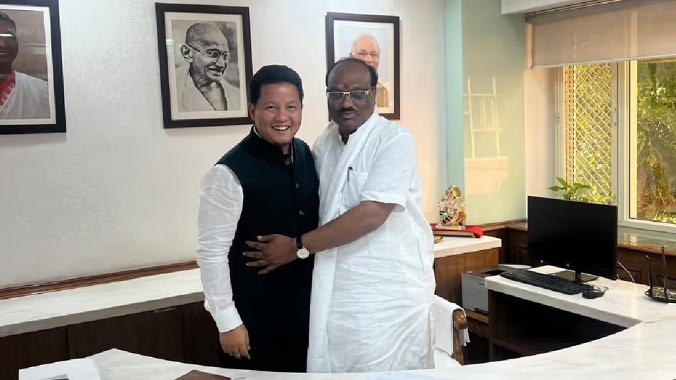 Durga Das Uikey, in a white kurta, embracing a visiting Member of Parliament in a dark waistcoat in his office, with framed portraits on the wall behind