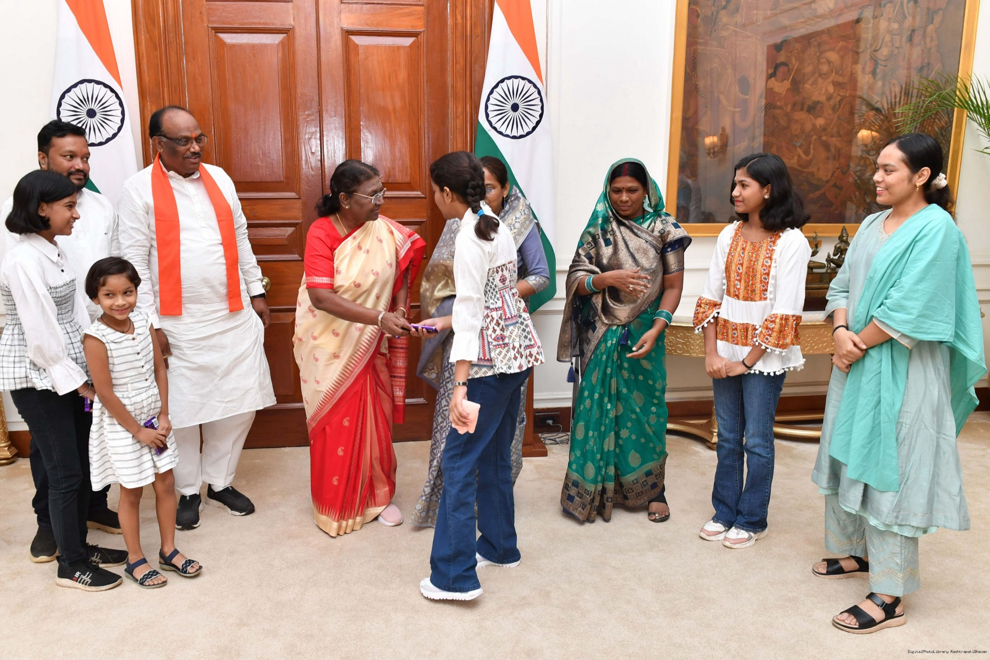 Durga Das Uikey, in a white kurta and saffron stole, with President Droupadi Murmu and a group of women and children in a hall at Rashtrapati Bhavan